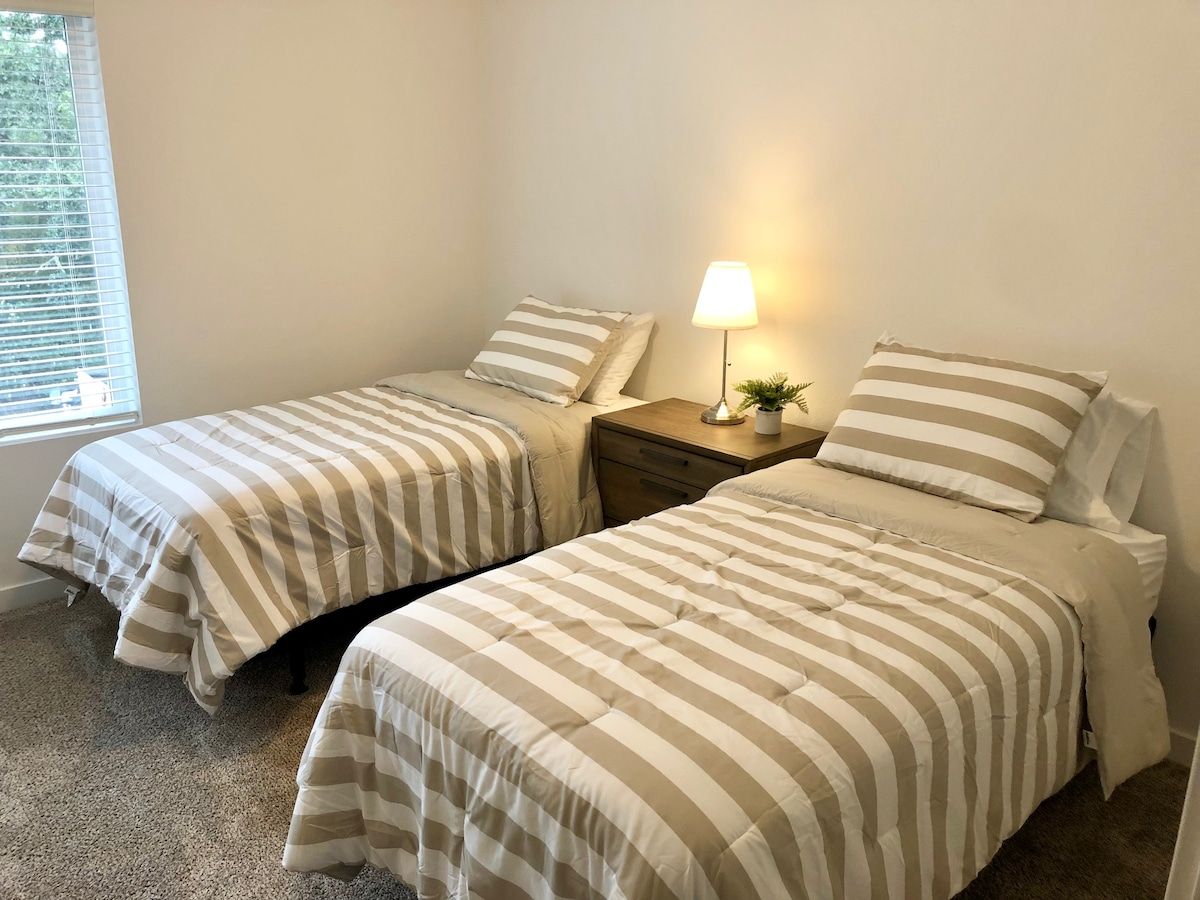Een slaapkamer met twee bedden, elk voorzien van gestreept beddengoed, een nachtkastje met een lamp en een raam met zonwering.