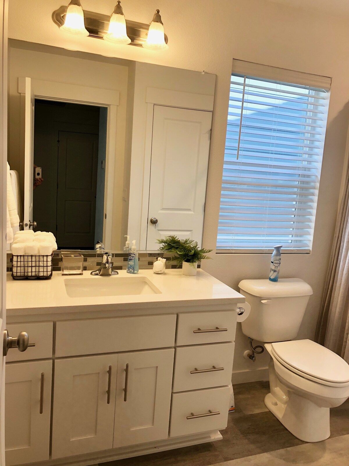 Une salle de bain avec un meuble-lavabo blanc, un miroir, des toilettes et une fenêtre avec des stores.
