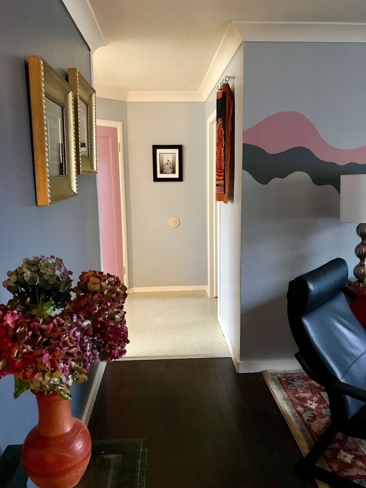 Un pasillo con una puerta, un cuadro en la pared, un jarrón con flores y una silla.