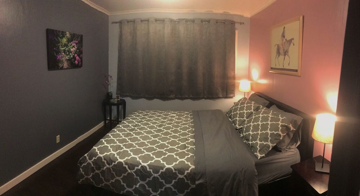 A bedroom with a bed, two bedside lamps, a window with a curtain, and a wall-mounted picture.