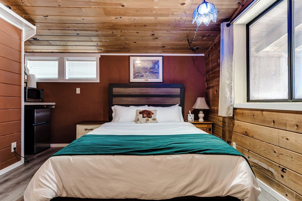 Een slaapkamer met een houten plafond, een bed met een groene dekbedovertrek, een nachtkastje met een lamp en een raam met jaloezieën.