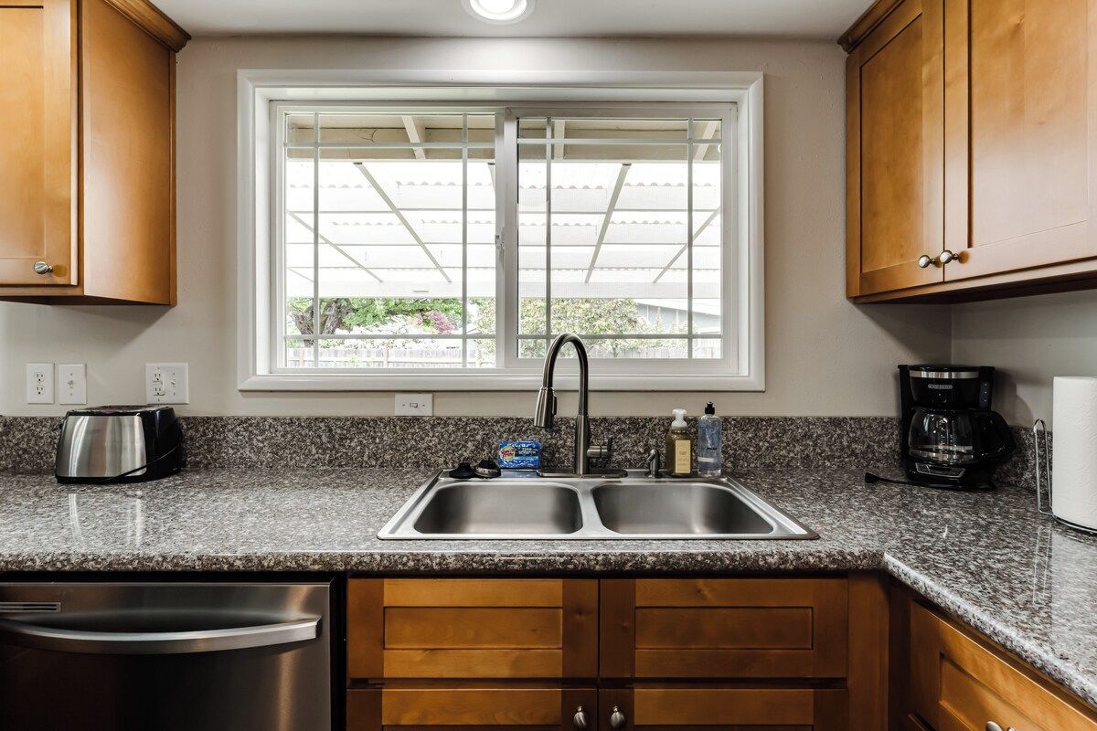 Cocina con encimeras de granito, electrodomésticos de acero inoxidable, armarios de madera y ventana con vista a la vegetación.