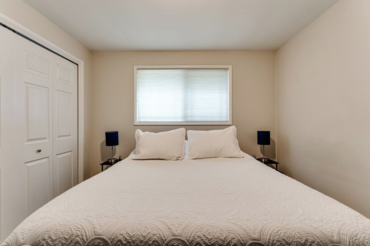 A bedroom with a large bed, two bedside tables with lamps, a window with blinds, and a door.