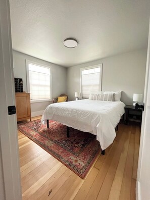 Een slaapkamer met een groot bed, een houten vloer, een rug met een patroon en twee ramen met zonwering.