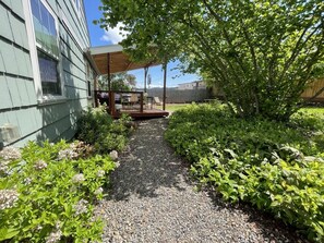 Ein Kiesweg führt zu einer Holzterrasse mit einer Pergola, umgeben von üppiger Vegetation und einem Gebäude mit heller Fassade.