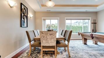 A dining area with a pool table, a dining table with chairs, and a view of the outdoors through large windows.