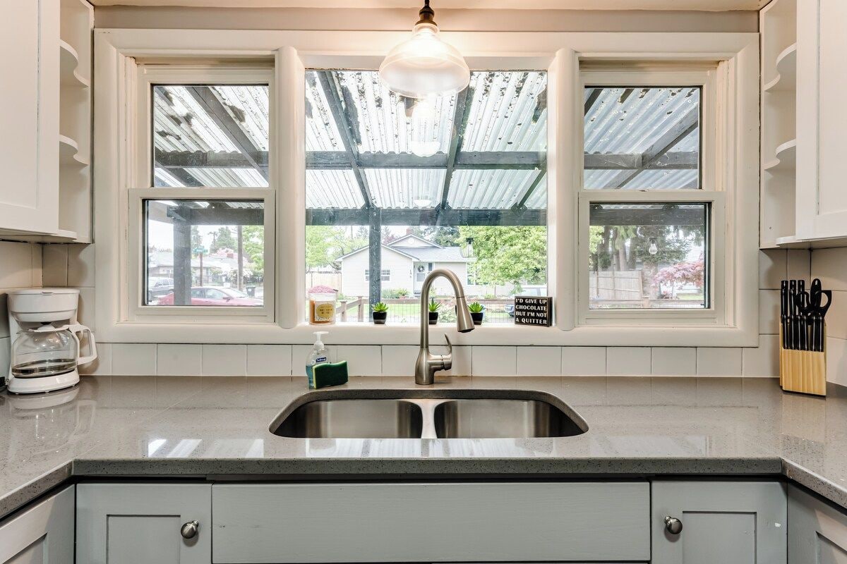 A kitchen with a double sink, a coffee maker, and a window with a view of a house and greenery.