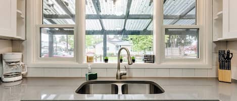 A kitchen with a double sink, a coffee maker, and a window with a view of a house and greenery.