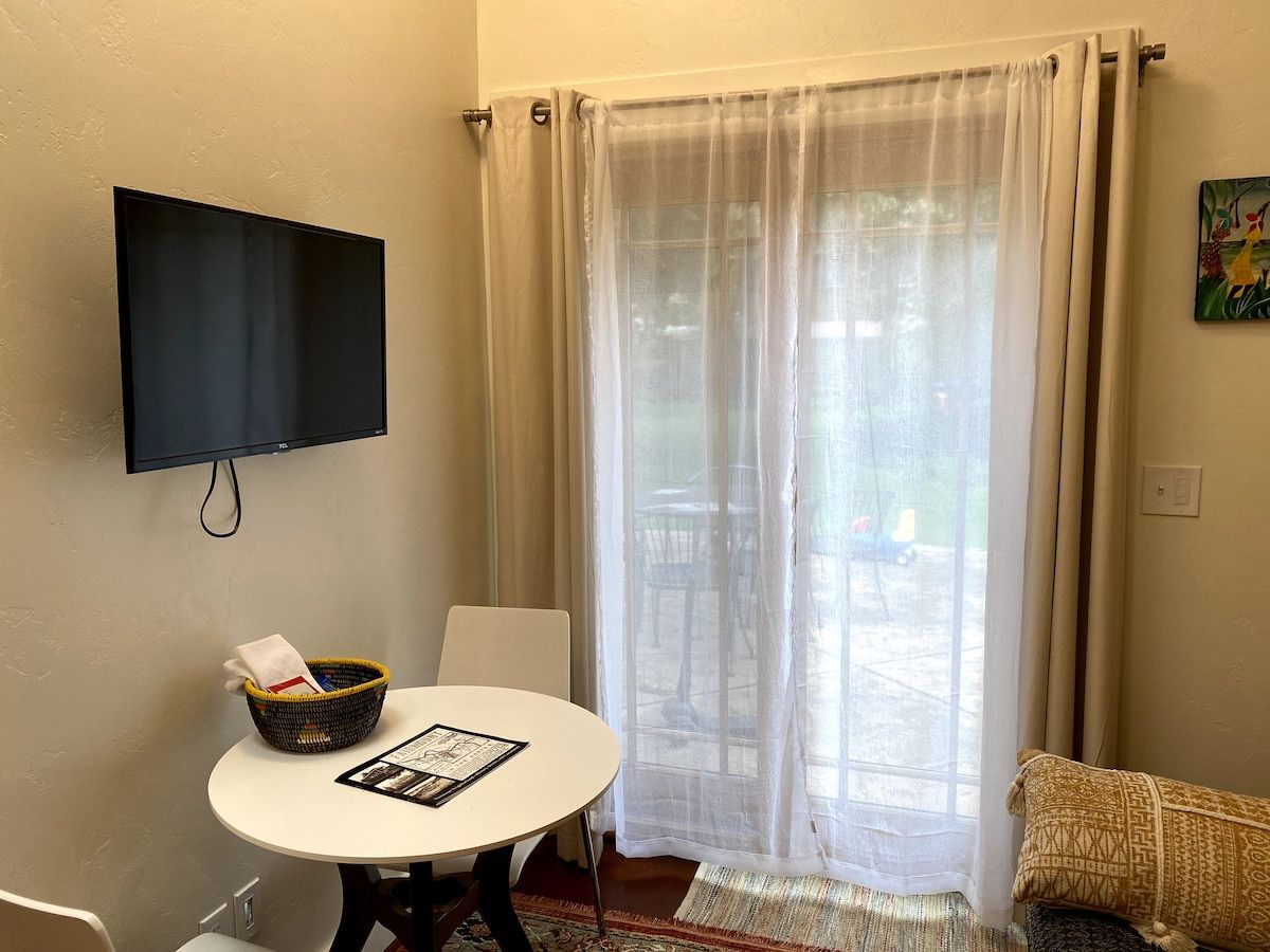 A room with a television mounted on the wall, a round table with a basket and a menu, and a sliding glass door leading to an outdoor area.