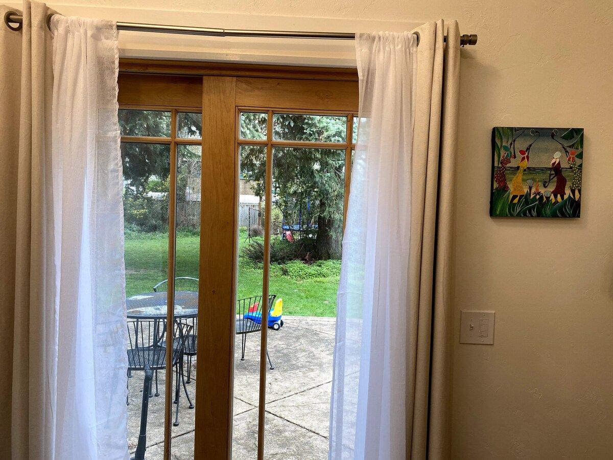 A view through a glass door with a white curtain, leading to an outdoor patio with a table and chairs, and a small toy car.