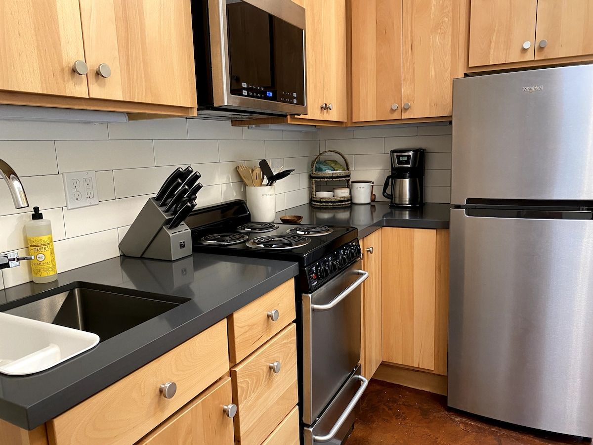 A modern kitchen with wooden cabinets, a stainless steel refrigerator, a black countertop, and a built-in oven.