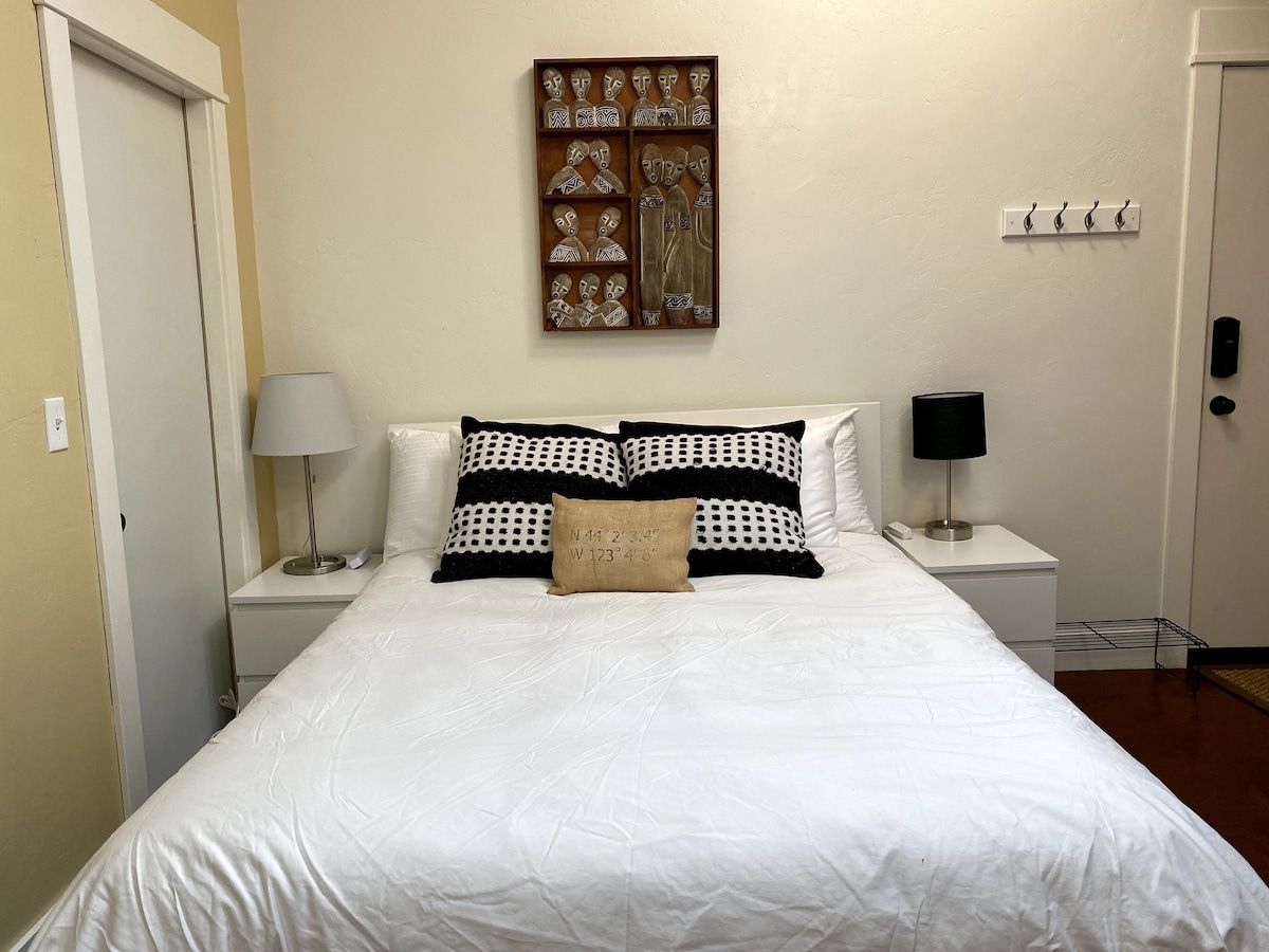 A neatly made bed with a white comforter and patterned pillows, a wall-mounted artwork, and a bedside table with a lamp.