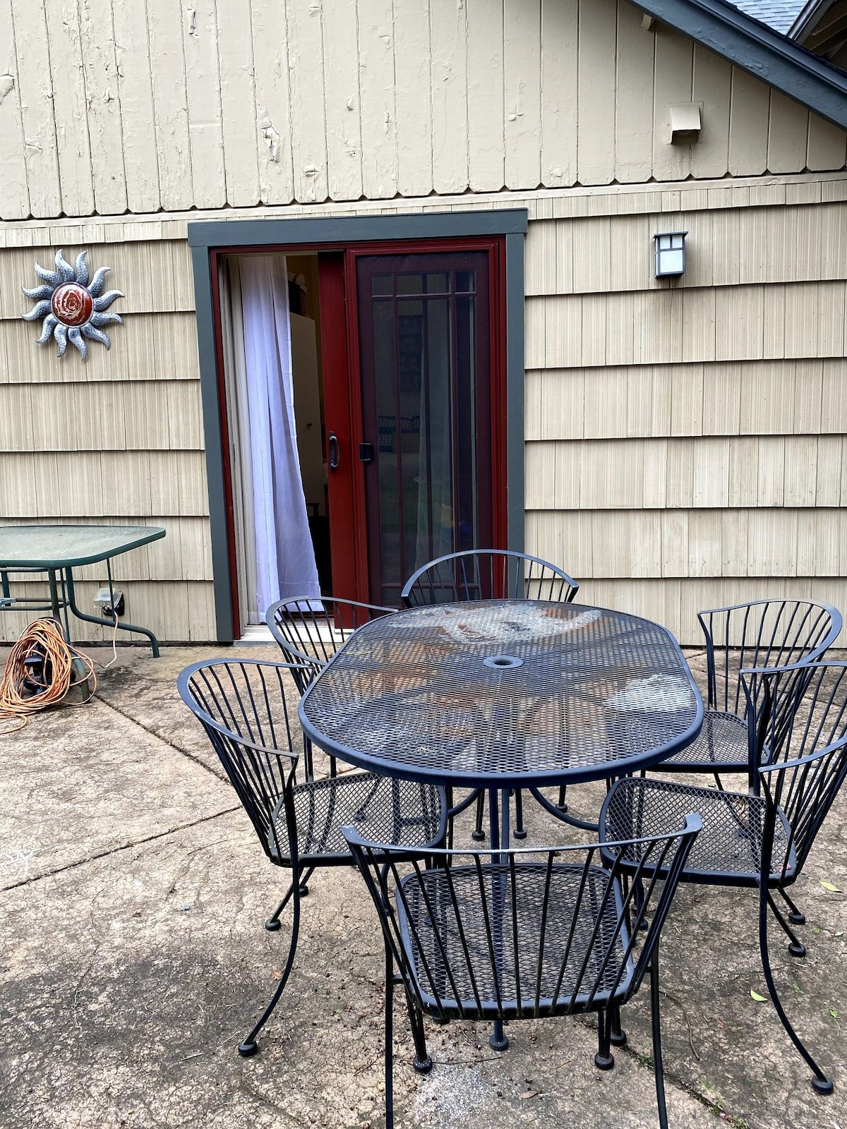 Eine Terrasse mit einem runden Metalltisch und Stühlen, einer dekorativen Sonne an der Wand und einer Tür mit rotem Rahmen.
