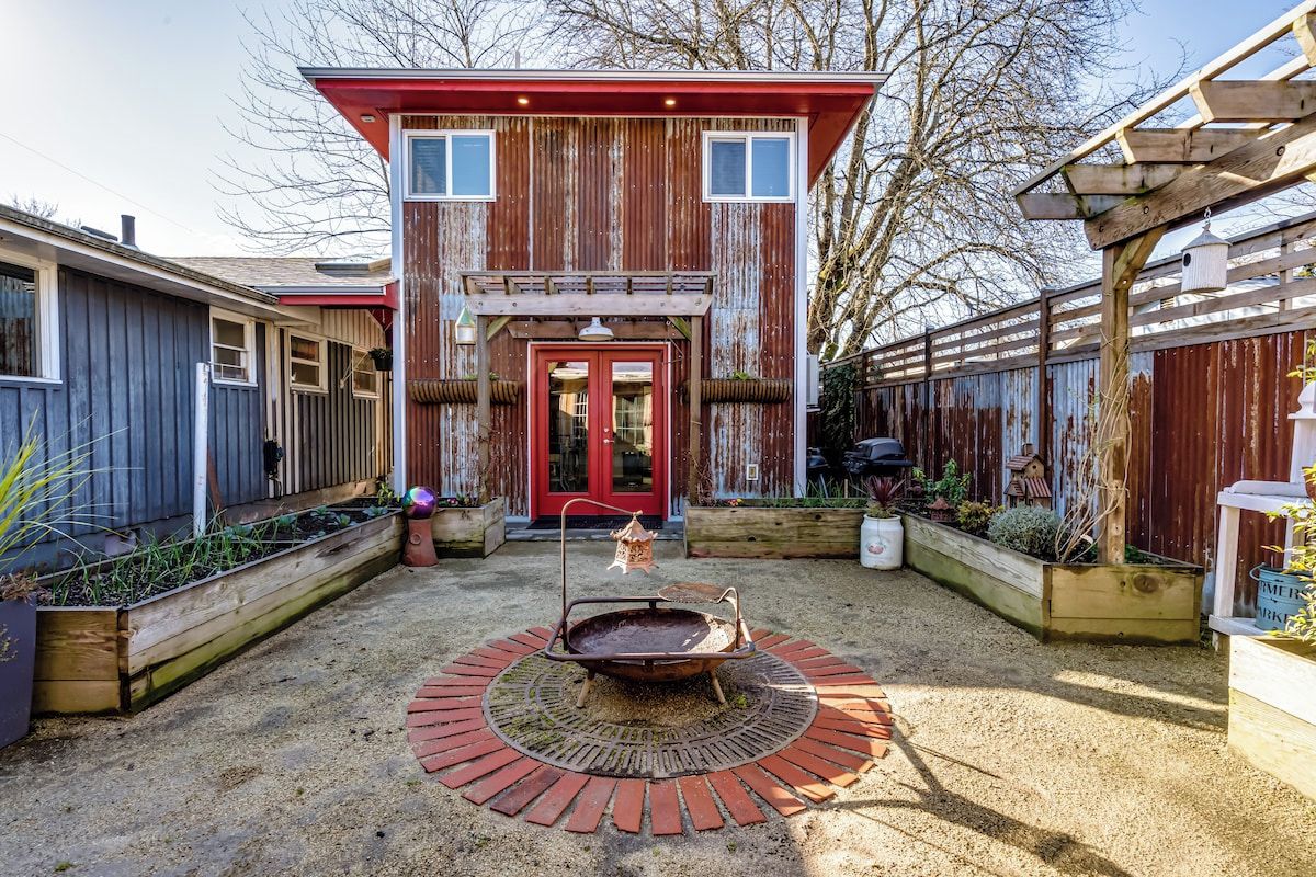 A rustic building with a red door, a wooden deck, and a circular fire pit in the center.