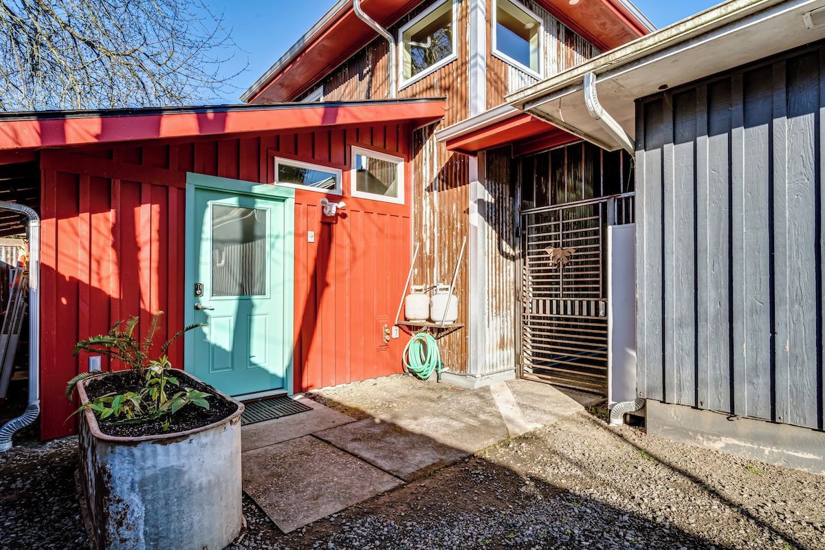 Un bâtiment aux couleurs rouge et grise, avec une porte bleue, un portail en métal et un petit jardin.