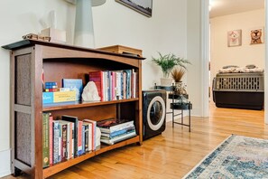 A room with a wooden bookshelf filled with books, a washing machine, a small table with decorative items, and a blue patterned rug. - Pet-Friendly Cottage in Walkable Whiteaker w/ (Eugene)