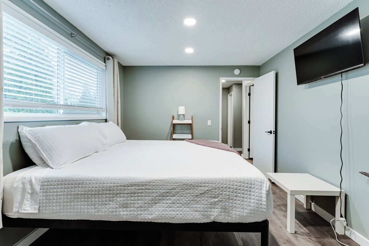 A bedroom with a bed, a bedside table, a TV mounted on the wall, and a window with blinds.
