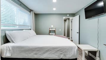 A bedroom with a bed, a bedside table, a TV mounted on the wall, and a window with blinds.