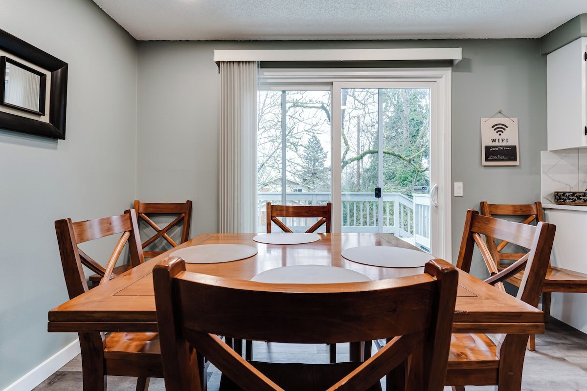 Un coin repas avec une table et des chaises en bois, une grande fenêtre donnant sur les arbres et un panneau indiquant la présence d’une connexion Wi-Fi sur le mur.