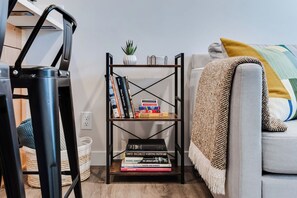A bookshelf with books and a small plant, a grey sofa with a throw blanket, and a black metal chair. - Dog-Friendly Studio w/ Patio & Parking, Walk to (Eugene)