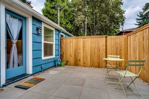 Ein Garten mit Holzzaun, einem blauen Haus, einer kleinen Terrasse mit Tisch und Stühlen und einem grünen Rasensprenger.