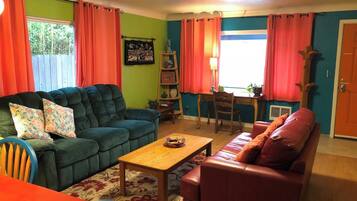 A living room with a sofa, a coffee table, and a red armchair. There is a window with curtains, a painting on the wall, and a wooden chair.