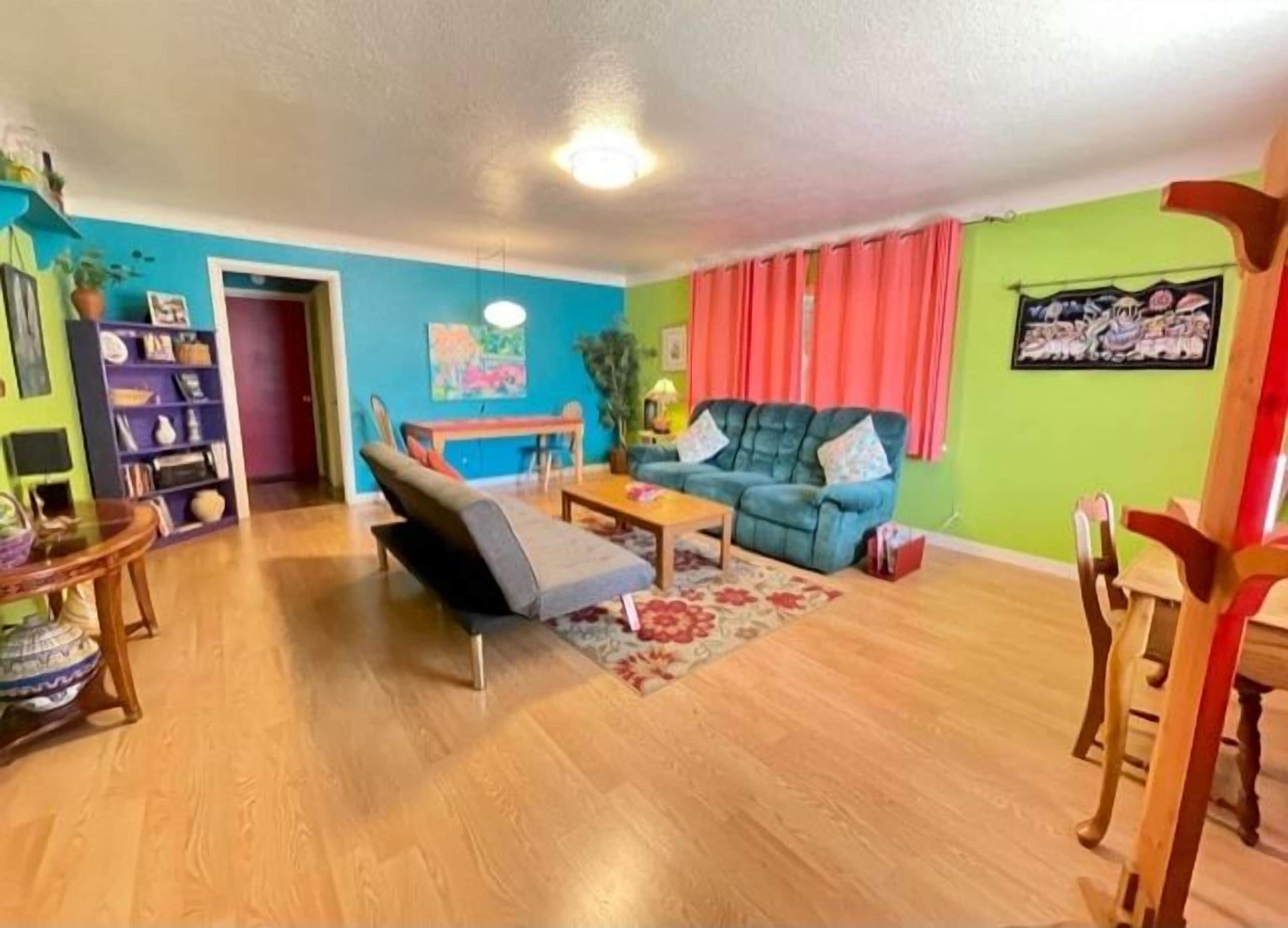 A living room with a sofa, a coffee table, and a dining table. There is a bookshelf and a door leading to another room.