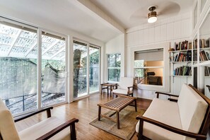 A spacious living room with a large sliding glass door leading to a garden, a wooden coffee table, a sofa, and a bookshelf filled with books. - Secluded Garden Retreat w/ Game Room & BBQ Patio (Eugene)
