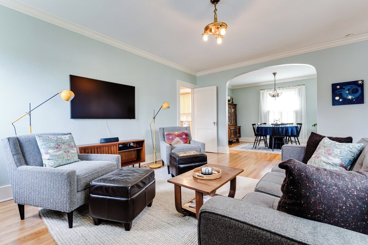 A living room with a sofa, armchair, ottoman, and a coffee table. There is a dining area with a table and chairs visible in the background.