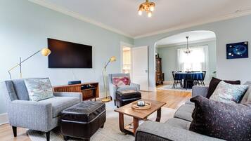 A living room with a sofa, armchair, ottoman, and a coffee table. There is a dining area with a table and chairs visible in the background.