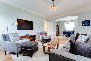 Ein Wohnzimmer mit einer Couch, Sesseln, Hocker und einem Couchtisch. Im Hintergrund ist ein Essbereich mit Tisch und Stühlen zu sehen.