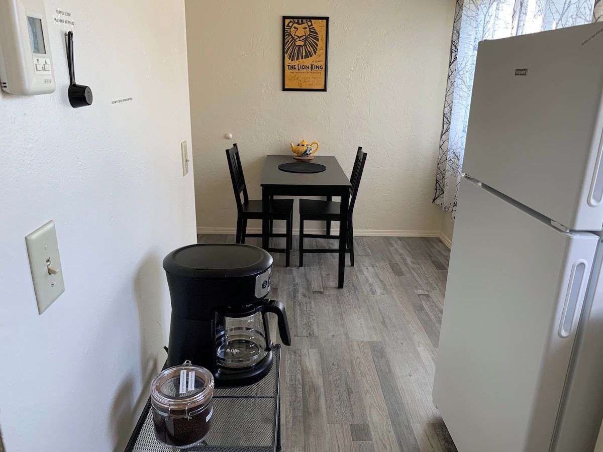 A compact kitchen with a coffee maker, a kettle, a refrigerator, and a framed picture on the wall.