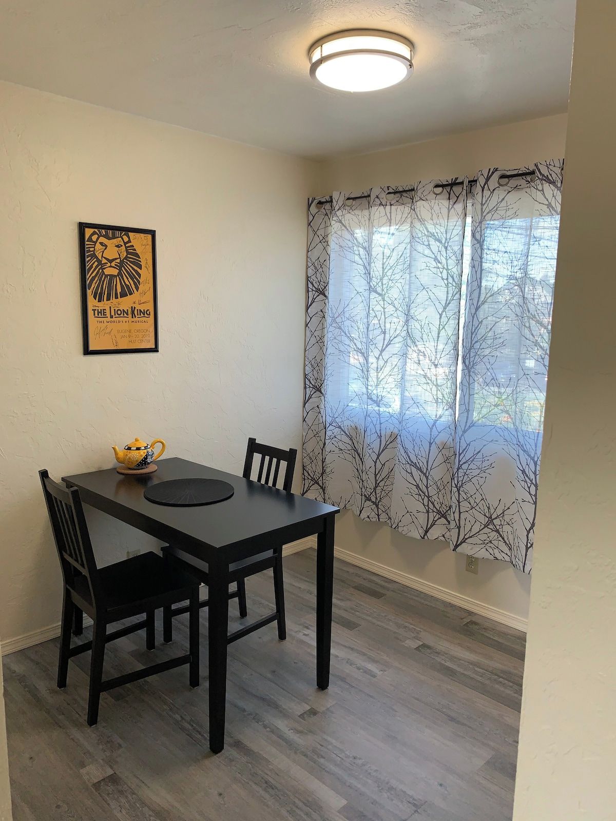 A dining area with a table, chairs, a framed poster, a window with a tree pattern curtain, and a ceiling light.