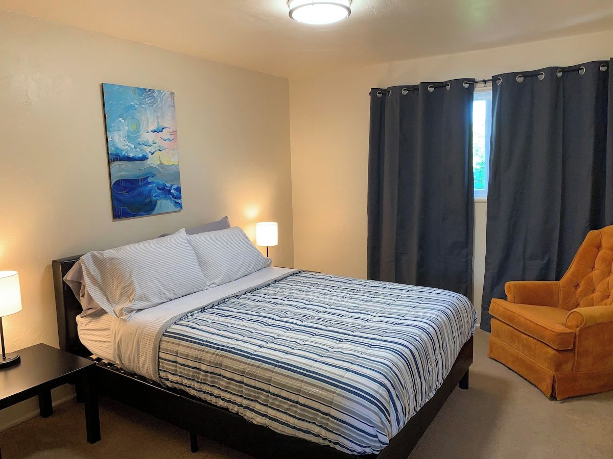 Quarto com cama, cadeira, quadro e janela com cortinas.
