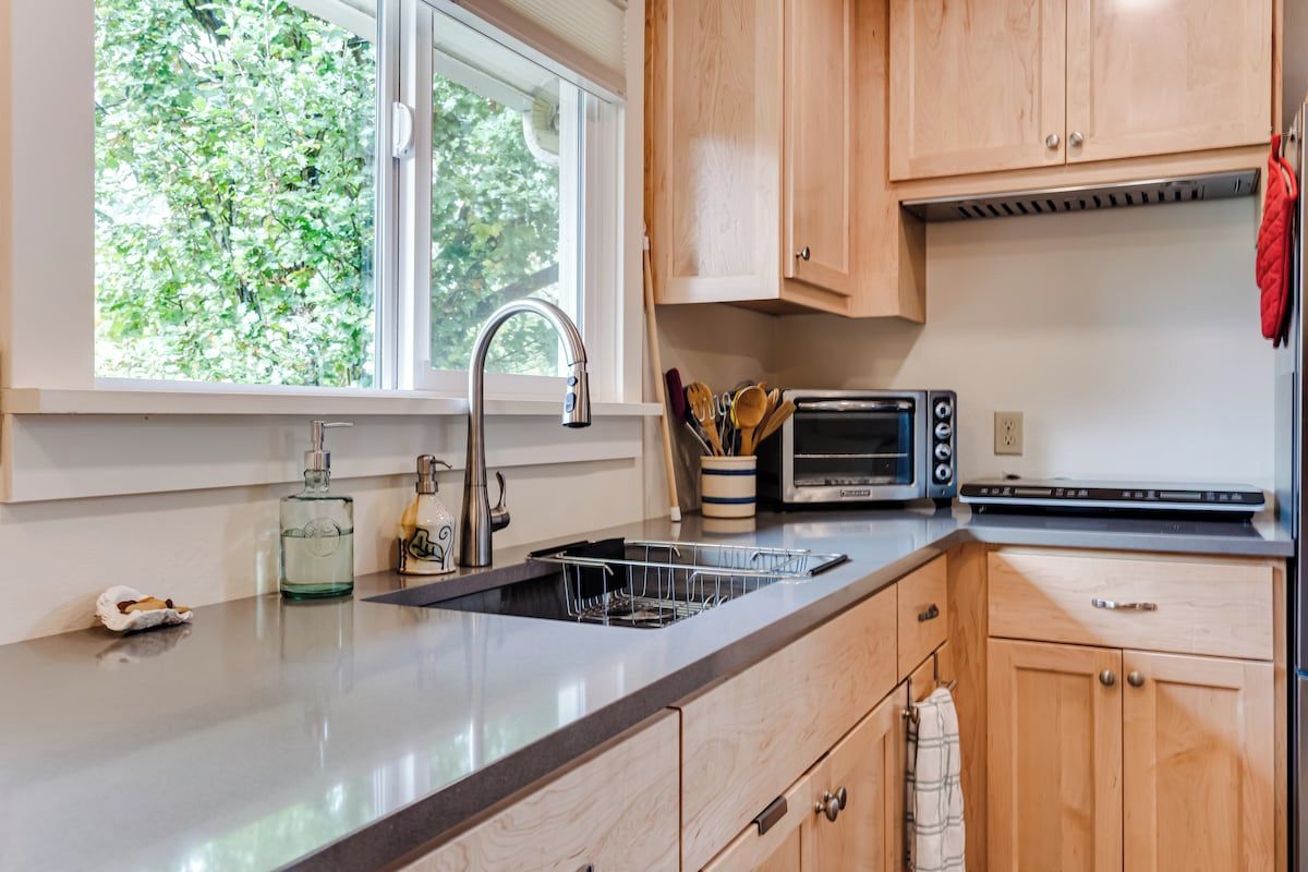 A kitchen with wooden cabinets, a stainless steel sink, a toaster, and a window with a view of trees.