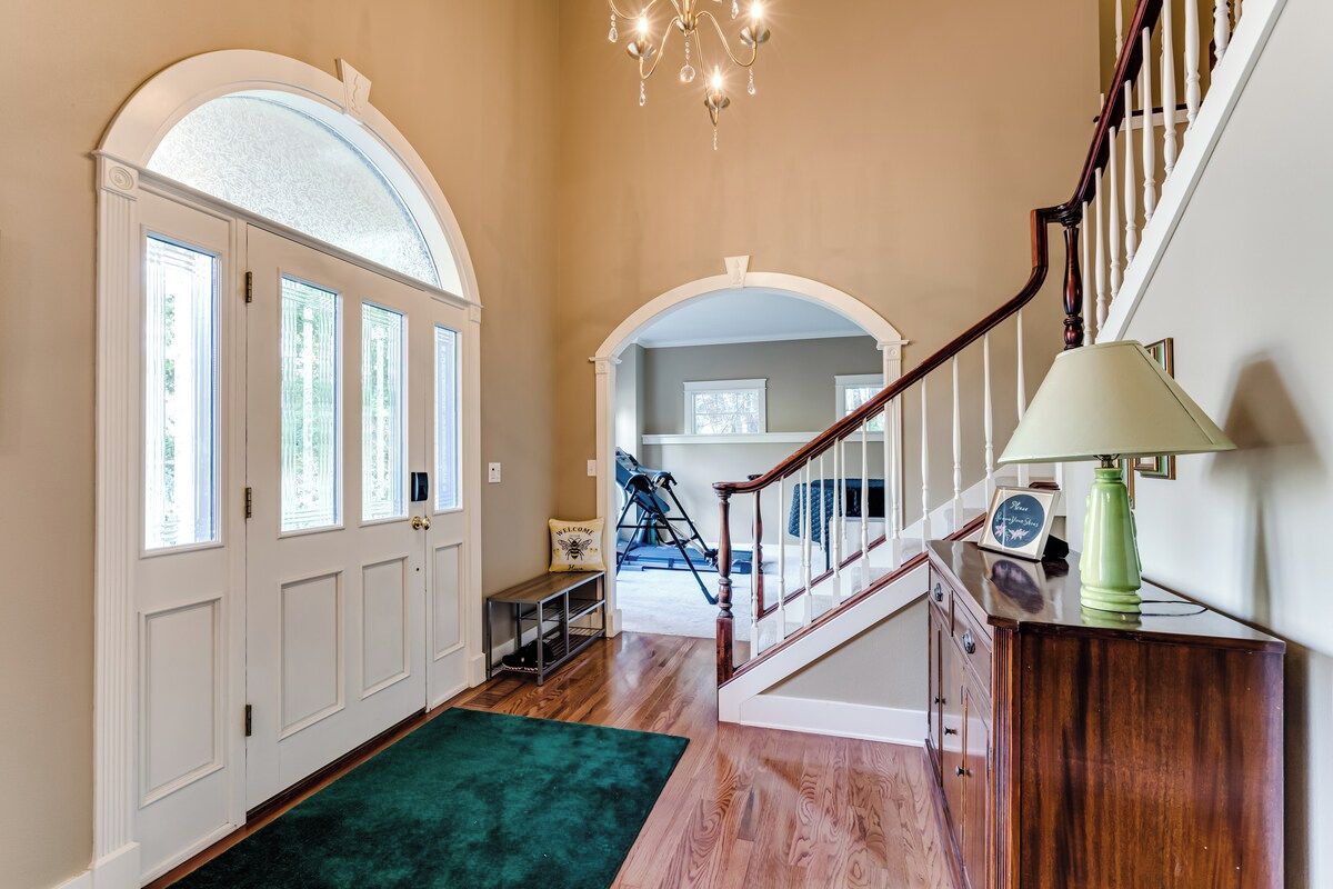 A hallway with a wooden floor, a green carpet, a staircase with a wooden railing, and a chandelier.