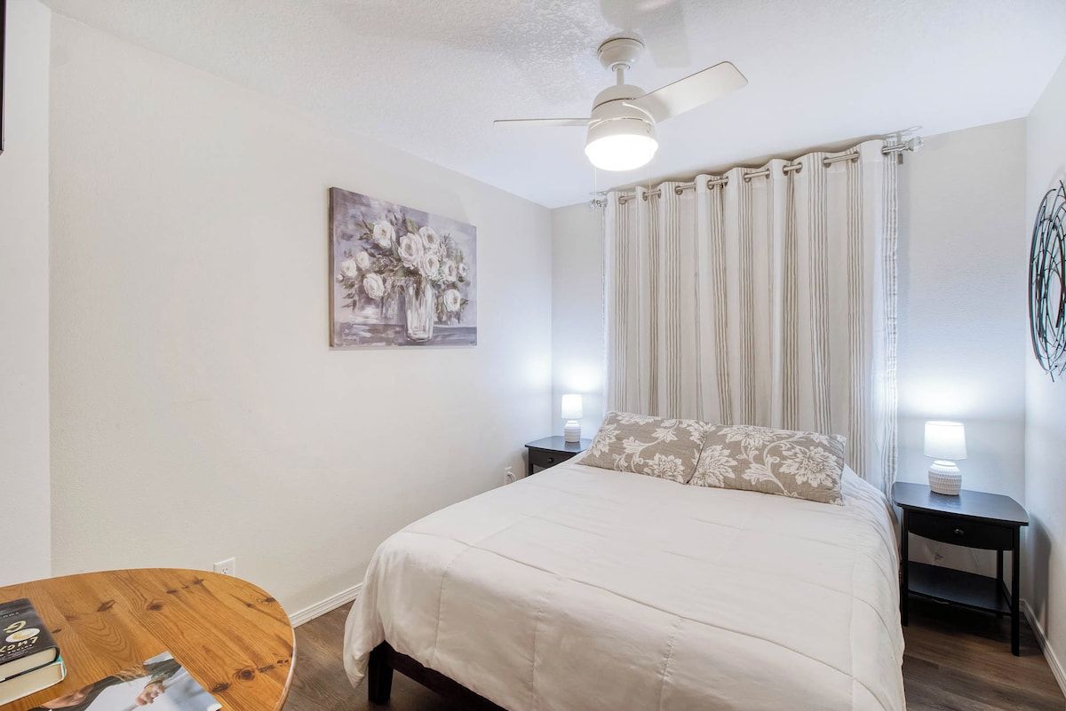A bedroom with a bed, nightstands, a ceiling fan, a painting, and curtains.