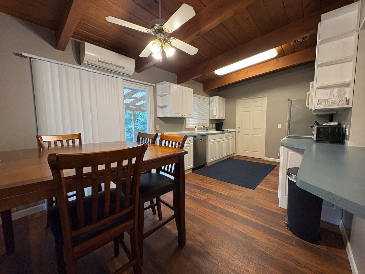 A kitchen with wooden flooring, white cabinets, a dining table with chairs, a ceiling fan, and a window with blinds.