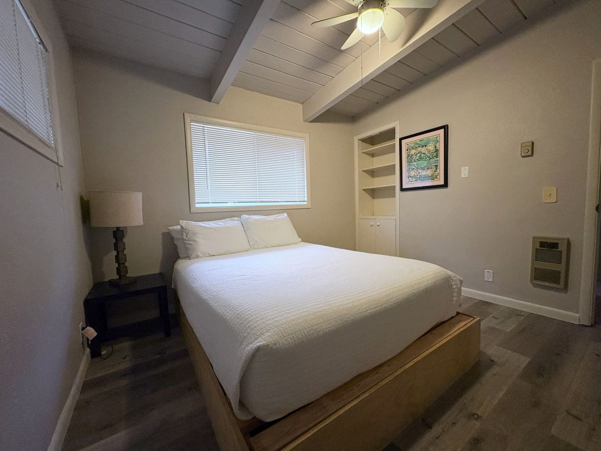 A bedroom with a bed, a nightstand, a lamp, a window with blinds, a ceiling fan, and a wall-mounted shelf with a framed picture.
