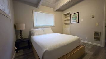 A bedroom with a bed, a nightstand, a lamp, a window with blinds, a ceiling fan, and a wall-mounted shelf with a framed picture.