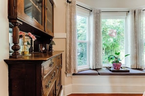 Une pièce avec un bow-window, une commode en bois, un vase rempli de fleurs et une plante en pot posée sur le rebord de la fenêtre.