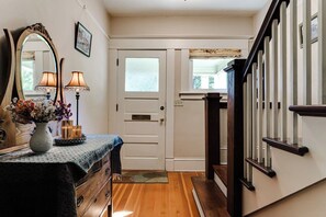 Un couloir avec un plancher en bois, une porte blanche, un miroir, un vase rempli de fleurs et une lampe.