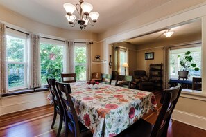 A dining room with a floral tablecloth, wooden chairs, a chandelier, and large windows with curtains. - Historic Manor w/ Hot Tub, Walk to UO & Hayward (Eugene)