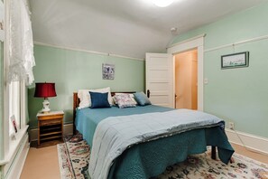 A bedroom with a bed, a nightstand, a lamp, a door, and a wall-mounted picture. - Historic Manor w/ Hot Tub, Walk to UO & Hayward (Eugene)