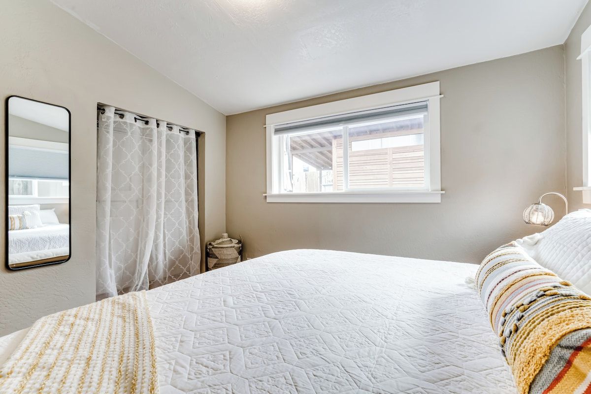 A bedroom with a large bed, a window with blinds, a mirror, and a bedside lamp.