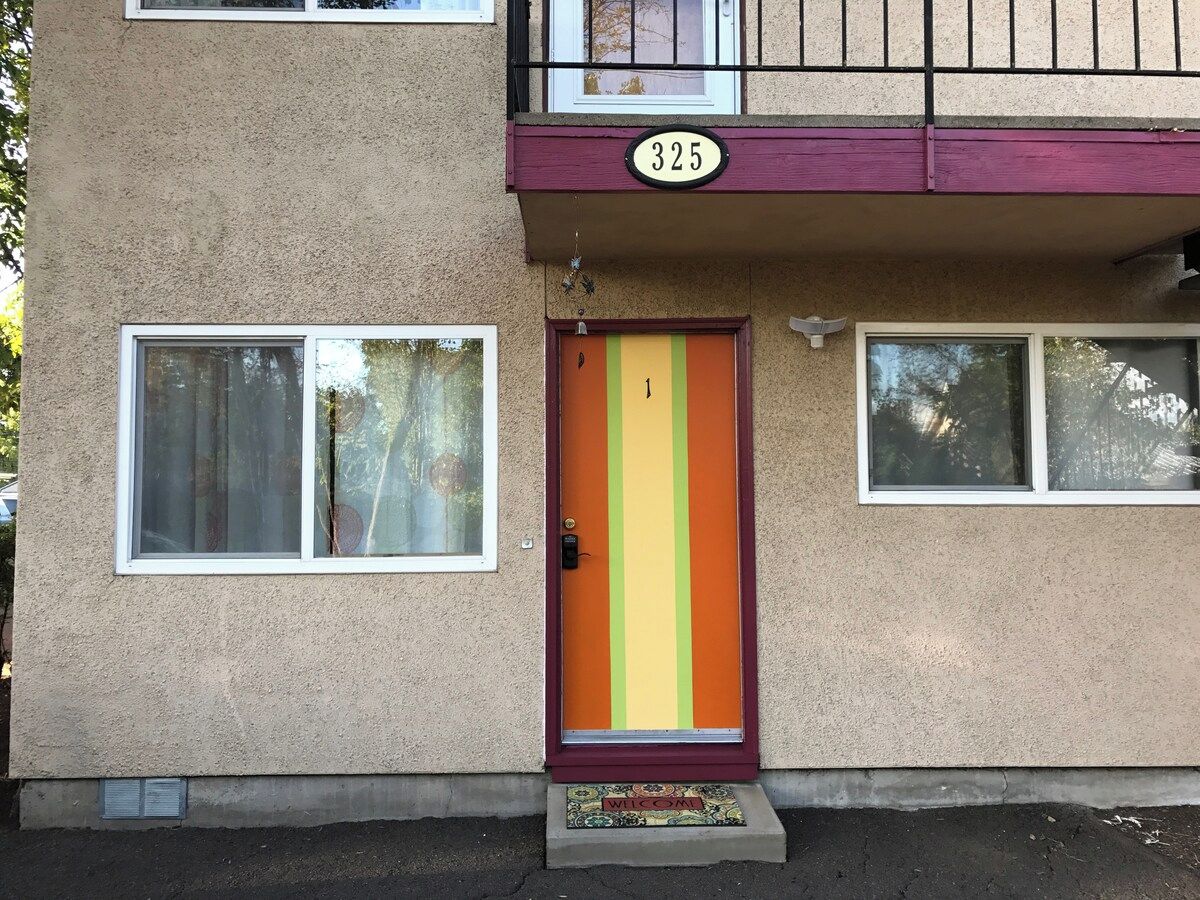 A two-story building with a beige exterior, a red-framed door with vertical stripes, and a balcony with a black railing.