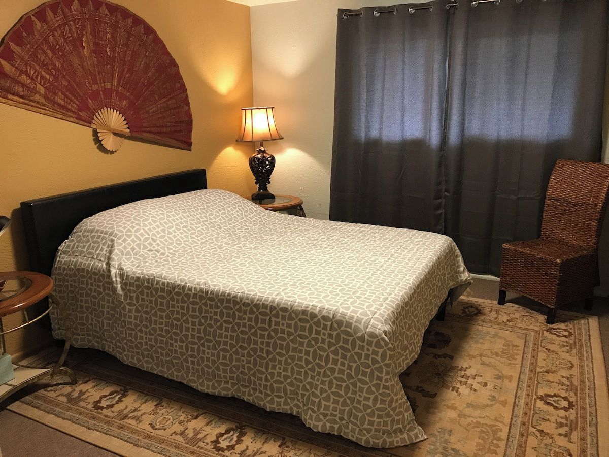A hotel room with a bed, a chair, a lamp, a wall fan, and a curtain.