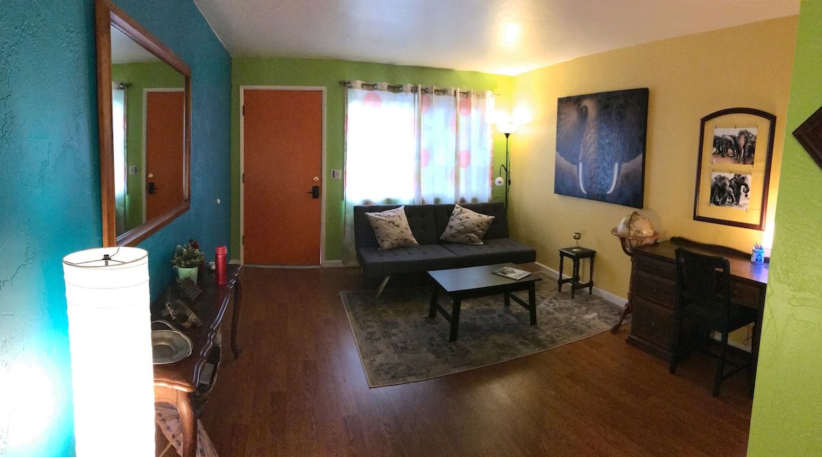 Sala de estar com sofá, mesa de centro e abajur. Há uma porta e uma janela com cortinas.