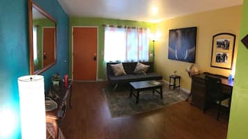 Sala de estar com sofá, mesa de centro e abajur. Há uma porta e uma janela com cortinas.