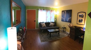 Sala de estar com sofá, mesa de centro e abajur. Há uma porta e uma janela com cortinas.